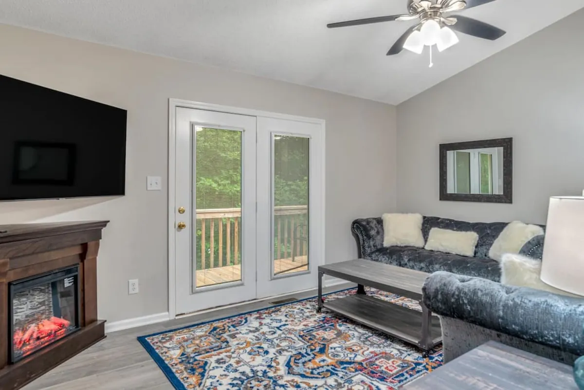 The image showcases a stylish, inviting living room with a plush sofa facing a modern fireplace, a soft area rug underfoot, and large sliding doors letting in natural light and hinting at outdoor space beyond. The atmosphere is cozy yet contemporary, perfect for both relaxation and gathering with friends or family.