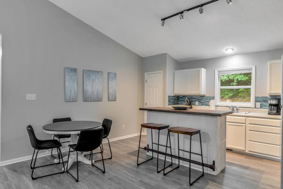 A bright, modern kitchen featuring a spacious island, stylish dining table, and vibrant wall artwork. The island is both a prep space and a gathering point, with light streaming in, hinting at a lively, social atmosphere. The arrangement suggests an easy transition between cooking and dining, perfect for connecting with others while preparing meals.