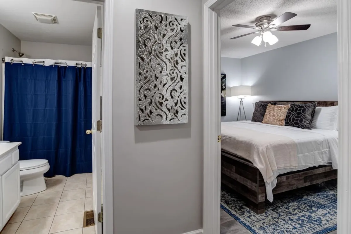 A cozy bedroom with a bed adorned with decorative pillows and ornate wall decor, adjacent to a bathroom with a blue shower curtain. The room exudes warmth and comfort, promising restful nights after lively outings.