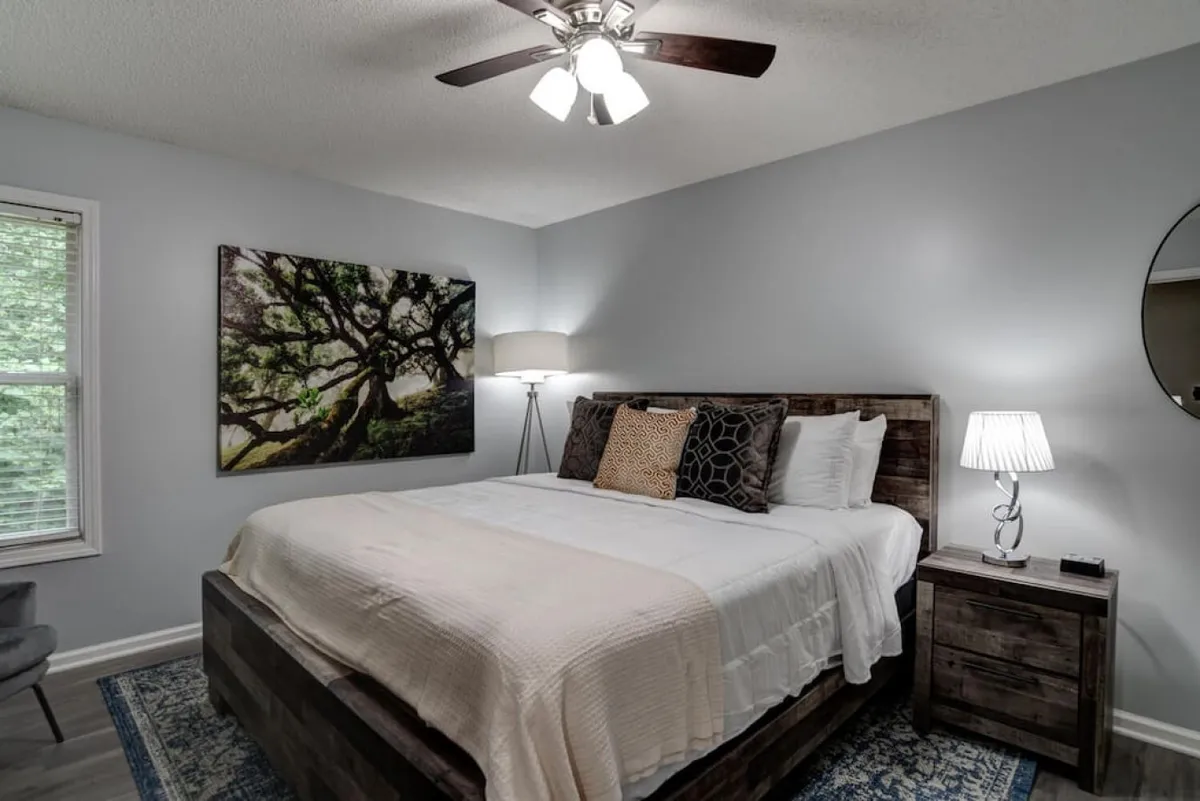 A cozy bedroom with a bed adorned with decorative pillows, a nightstand and lamp, and striking tree-themed artwork. Natural light filters softly through a window, contributing to the relaxing and serene mood of the space.