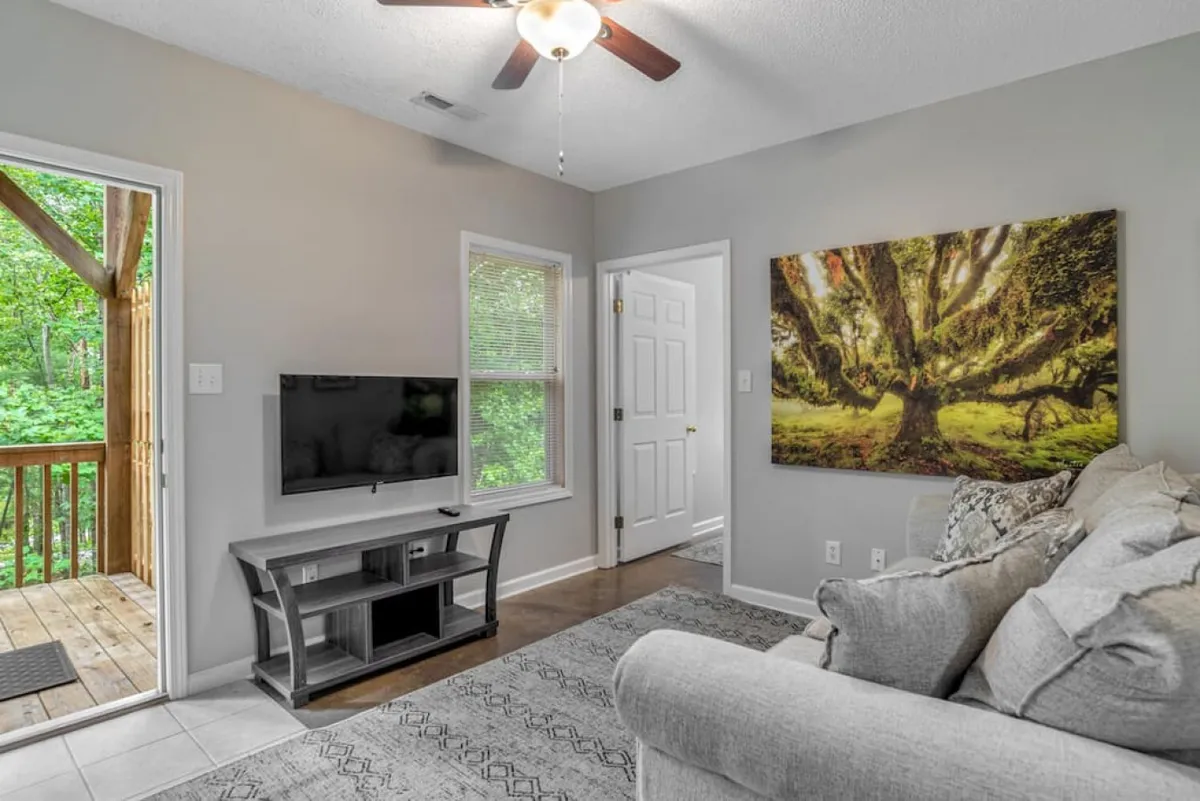 This image features a comfortable sofa, large artwork, a television, and an open door leading out to the deck. The space is bright with natural light, and the layout suggests an inviting atmosphere where people can gather, watch TV, and socialize. The sightline to the outdoors adds a sense of openness and connection.