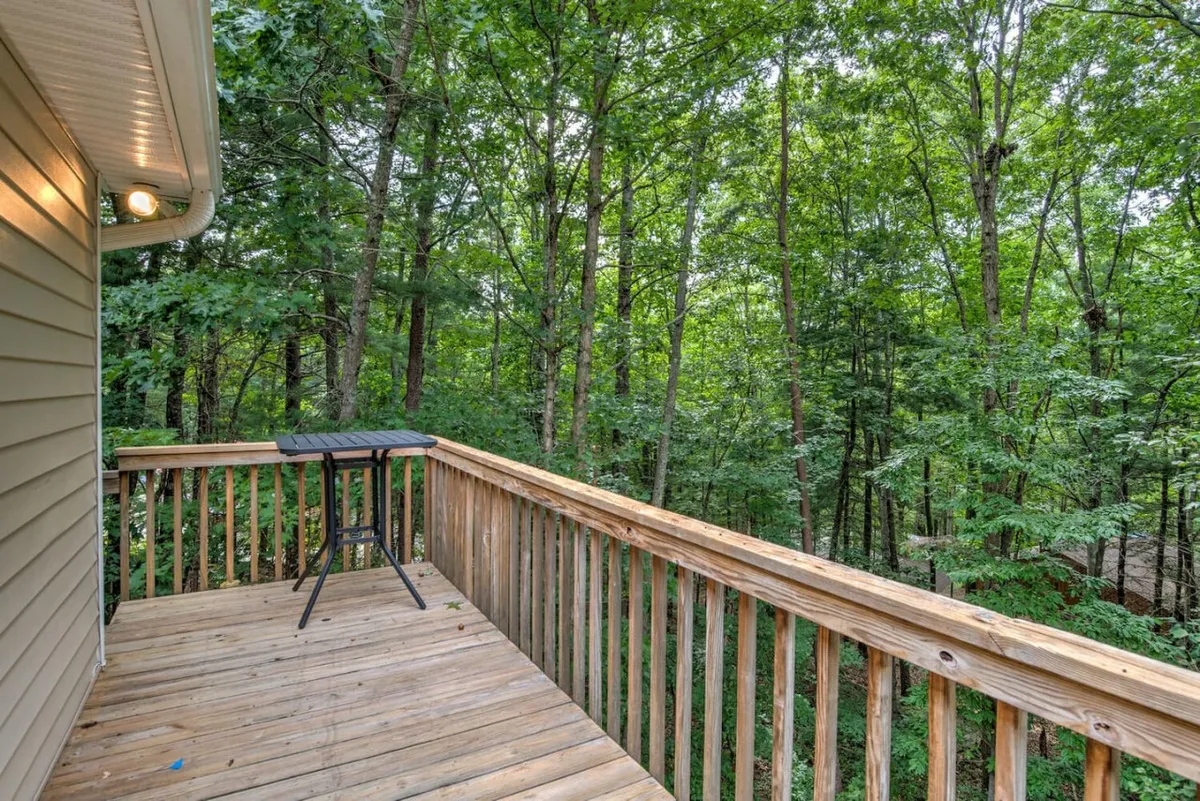 This exterior image showcases a wooden deck with a sturdy railing surrounded by lush trees. A small table is set for relaxing or enjoying a morning coffee, with natural sunlight filtering through the foliage, creating a tranquil and welcoming atmosphere.